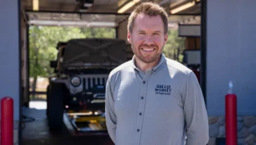 A man in a light gray Grease Monkey shirt stands smiling outside an auto repair shop, positioned near the primary navigation area of the lot, with a black Jeep and open garage in the background.