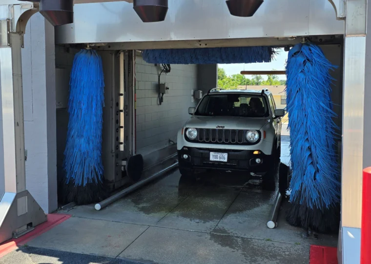 A Jeep is positioned inside an automatic car wash with blue brushes on either side, preparing to be cleaned.