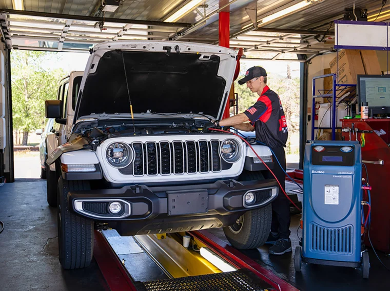 Auto mechanic in uniform works under the hood of a white SUV inside a garage, using diagnostic equipment connected to a nearby machine as part of comprehensive air conditioning repair services.
