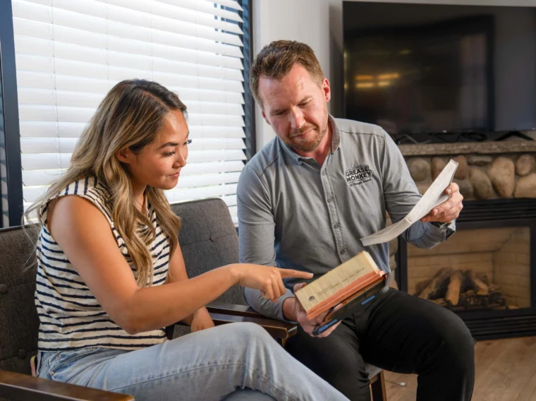 A woman and a man sit indoors; she points at a book while he holds a paper and looks at the book. They appear to be discussing something together, possibly reviewing details about filter services.