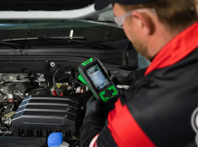 A mechanic holds a diagnostic scanner and checks a vehicle’s engine while wearing gloves, safety glasses, and a red and black uniform, ensuring expert Battery Services are part of the inspection.