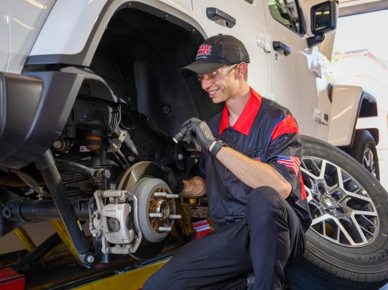 A mechanic wearing gloves and a uniform inspects a vehicle’s brake system while the car is lifted on a hydraulic lift, performing thorough Brake Services to ensure optimal safety and performance.