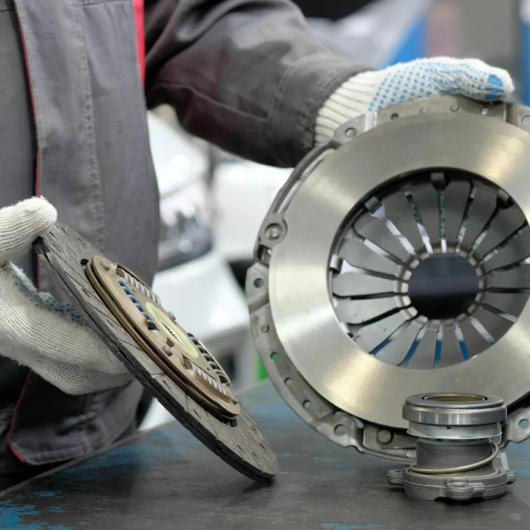 A person wearing gloves holds a car clutch disc and pressure plate assembly during a clutch replacement, with additional clutch components resting on a workbench.