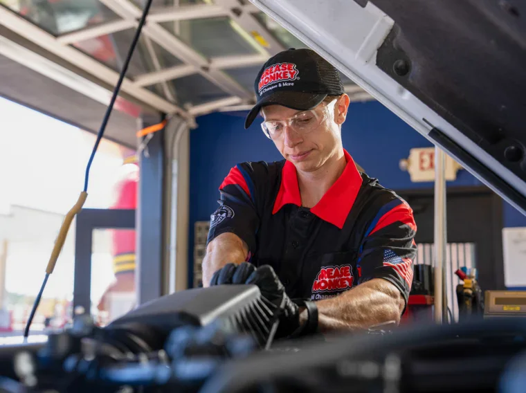 A mechanic wearing safety glasses and gloves works under the hood of a car in a garage, carrying out filter services, with tools and equipment visible in the background.