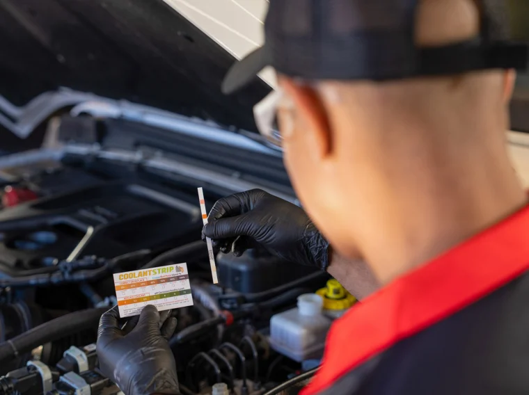 A person wearing gloves holds a test strip and instruction card while checking engine coolant quality beside an open car hood, highlighting the importance of regular maintenance for both engine performance and effective air conditioning.