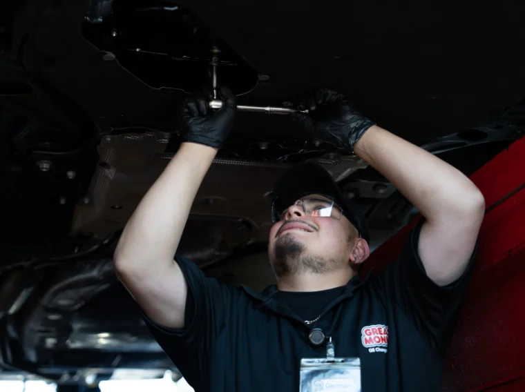 A mechanic wearing gloves and safety glasses works under a car in a garage, using a wrench to tighten or loosen a bolt.