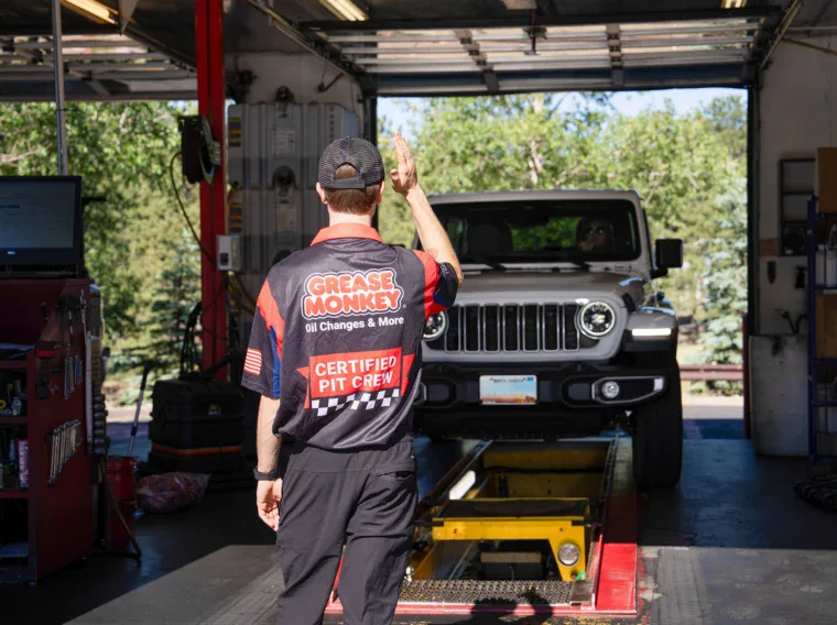 An auto technician signals while guiding a gray Jeep onto a lift inside a service garage. The technician, ready to assist with air conditioning services, wears a “Grease Monkey” uniform with “Certified Pit Crew” on the back.