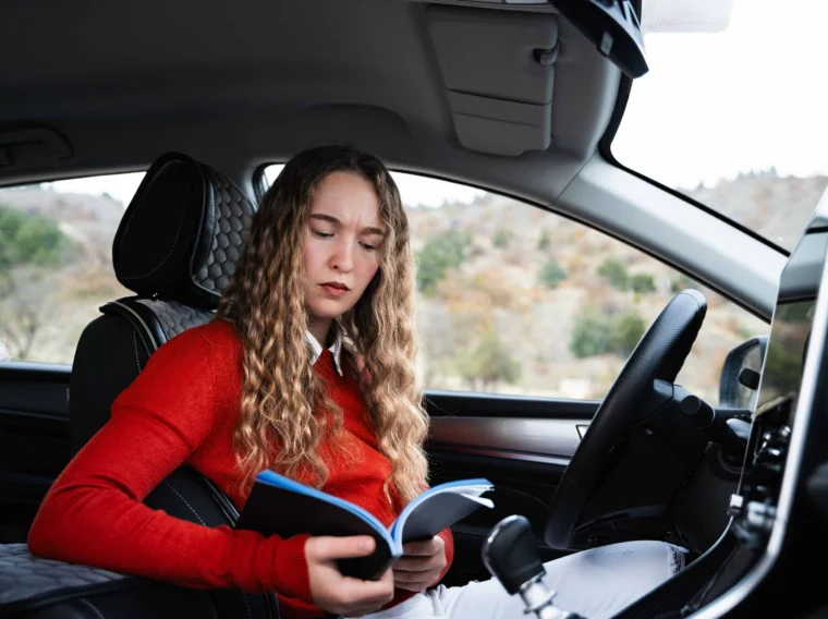 A woman with long curly hair reads a book while sitting in the driver's seat of a parked car, perhaps waiting for her vehicle's Filter Services to be completed.