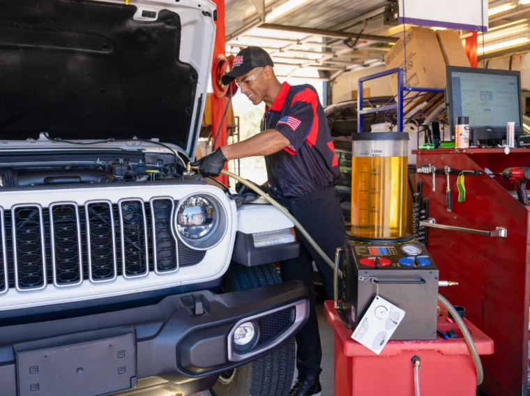 A mechanic in uniform works under the hood of a white SUV inside an auto repair shop, surrounded by diagnostic and fluid equipment.