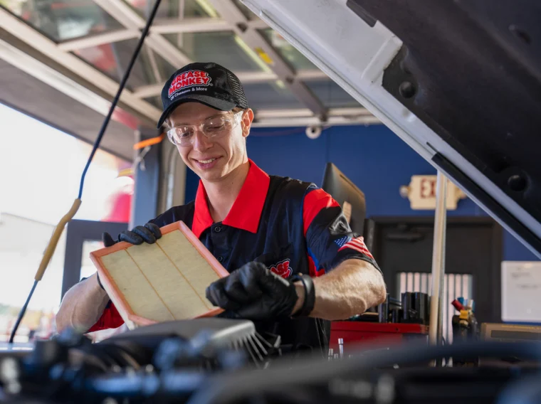 An auto mechanic in uniform and gloves inspects an air filter while working under the hood of a vehicle in a garage, demonstrating the importance of regular Filter Services for optimal engine performance.