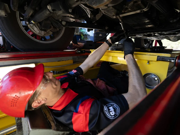 A mechanic wearing a red hard hat and gloves works underneath a vehicle, using a tool to make repairs in an auto service garage.