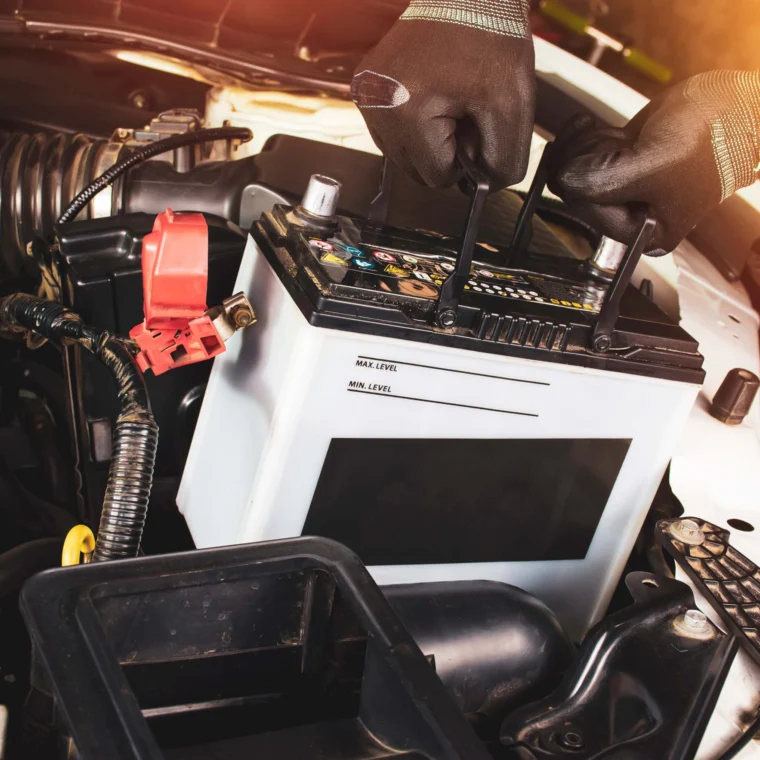 Two hands holding a car battery underneath the hood of a vehicle.