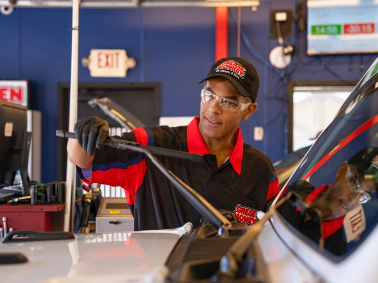 A mechanic wearing gloves and safety glasses inspects or performs a windshield wiper replacement on a vehicle inside an auto service shop.