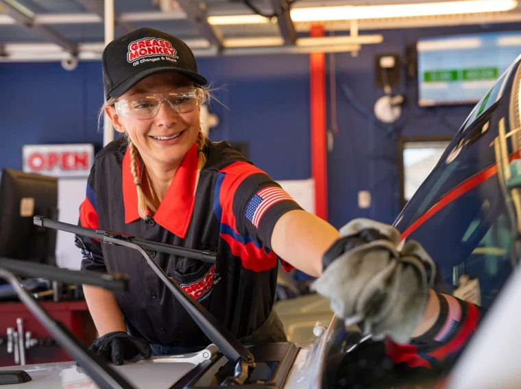 A mechanic in uniform and safety glasses cleans a car windshield inside a garage, with equipment, an "OPEN" sign, and wiper blades visible in the background—highlighting their dedication to quality wiper services.