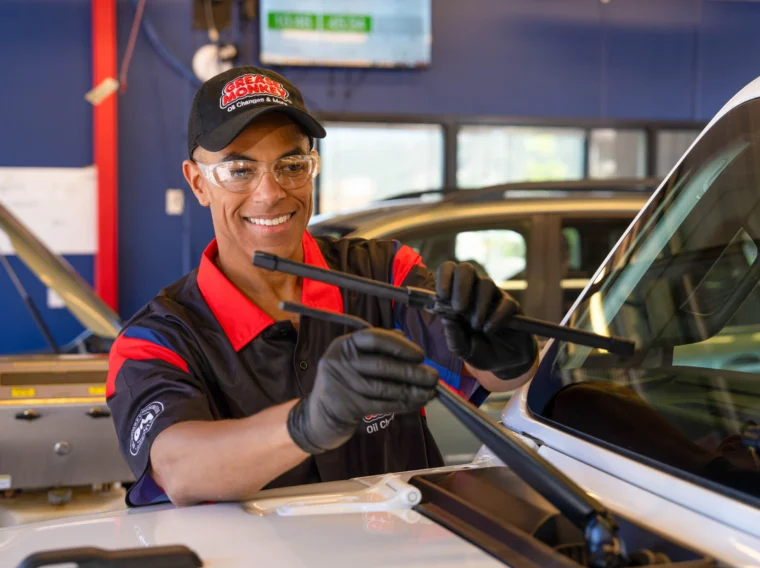 A technician wearing gloves and safety glasses is smiling while replacing a windshield wiper on a vehicle inside an auto service shop.