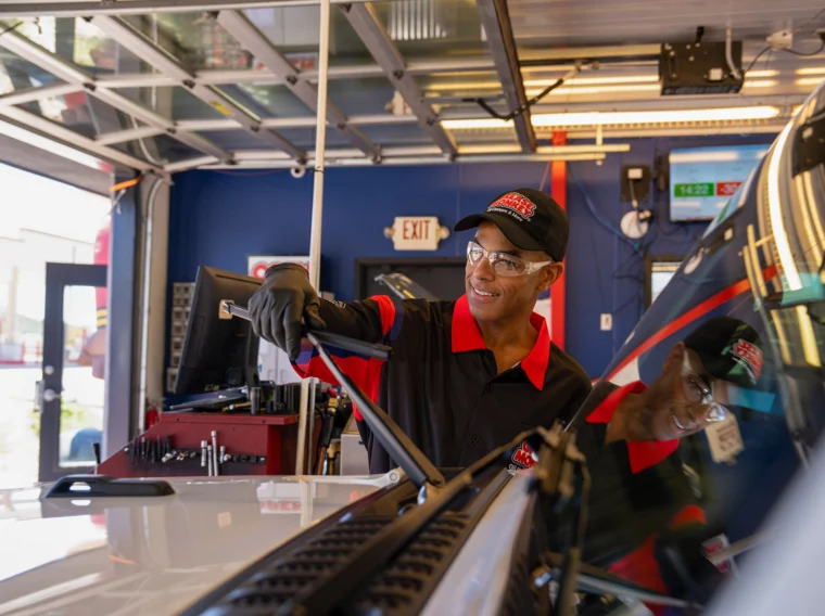 A mechanic wearing a uniform and cap performs wiper replacement by fitting a new wiper blade on a vehicle inside a well-lit automotive service garage.