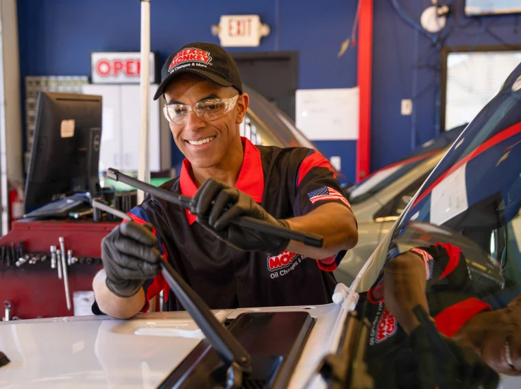 A mechanic wearing safety glasses and gloves performs a windshield wiper replacement, carefully installing new wipers on a car inside an auto service shop.