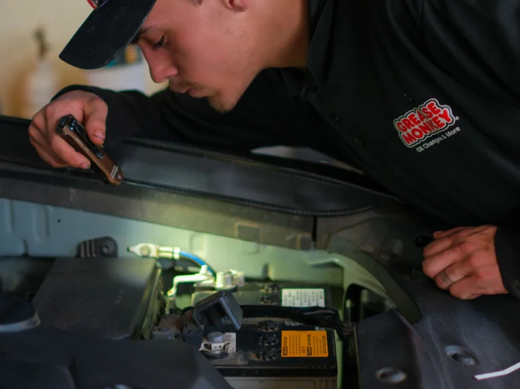 A mechanic in uniform inspects a car battery with a flashlight, leaning over the open hood in a workshop during an AC repair.