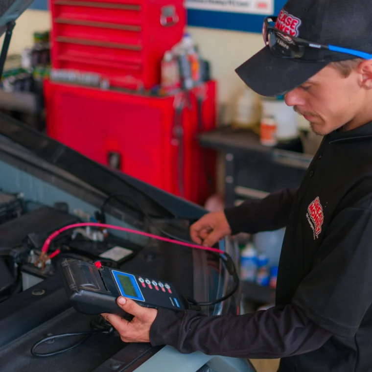A mechanic uses an electronic diagnostic tool to check a car battery while performing Battery Services in a garage with a red tool cabinet in the background.