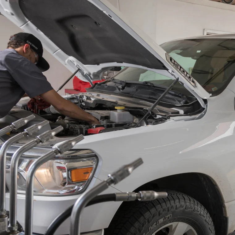 A mechanic inspects under the hood of a white Ram truck, possibly checking Battery Services, while a person sits inside the vehicle at an automotive repair shop.