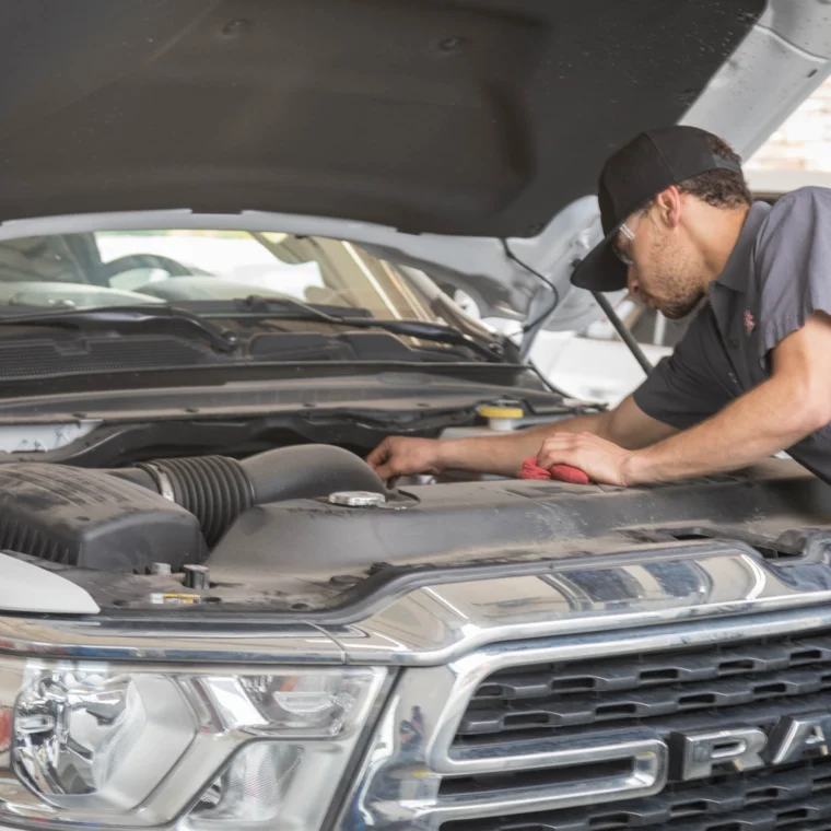 A mechanic wearing a hat and safety glasses inspects the engine of a white RAM truck with the hood open in a garage, performing routine checks and Battery Services.