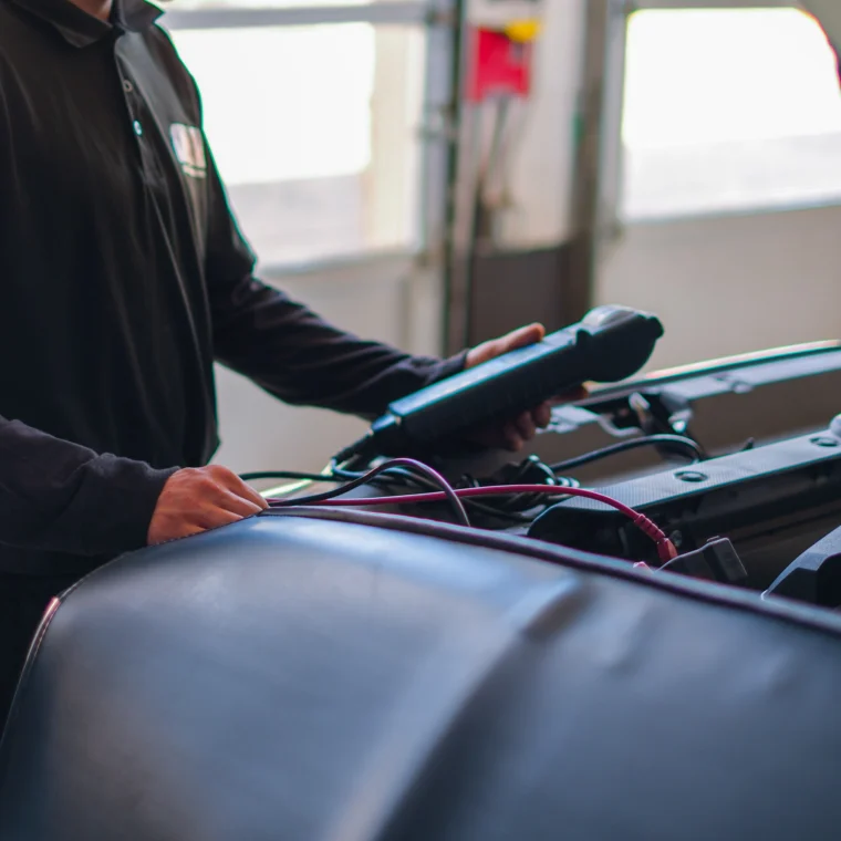 A mechanic wearing a cap uses a diagnostic tool under the hood of a car in a garage, checking essential components during battery services.