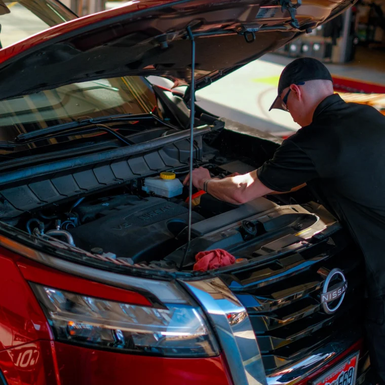 A mechanic works under the hood of a red Nissan SUV in a garage, performing battery services and essential maintenance or repairs.