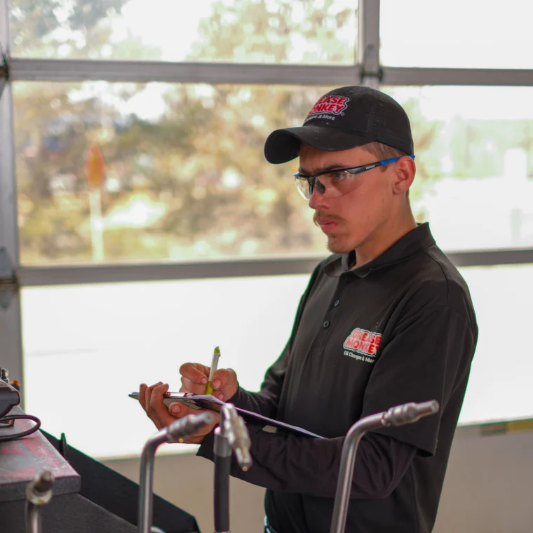 A man in a black Grease Monkey uniform and hat writes on a clipboard while standing near automotive equipment in a garage, preparing to perform expert brake services.