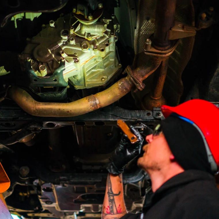 A mechanic wearing a red helmet and gloves inspects the underside of a vehicle, holding a flashlight and looking up at the car’s transmission, exhaust system, and brake services components.