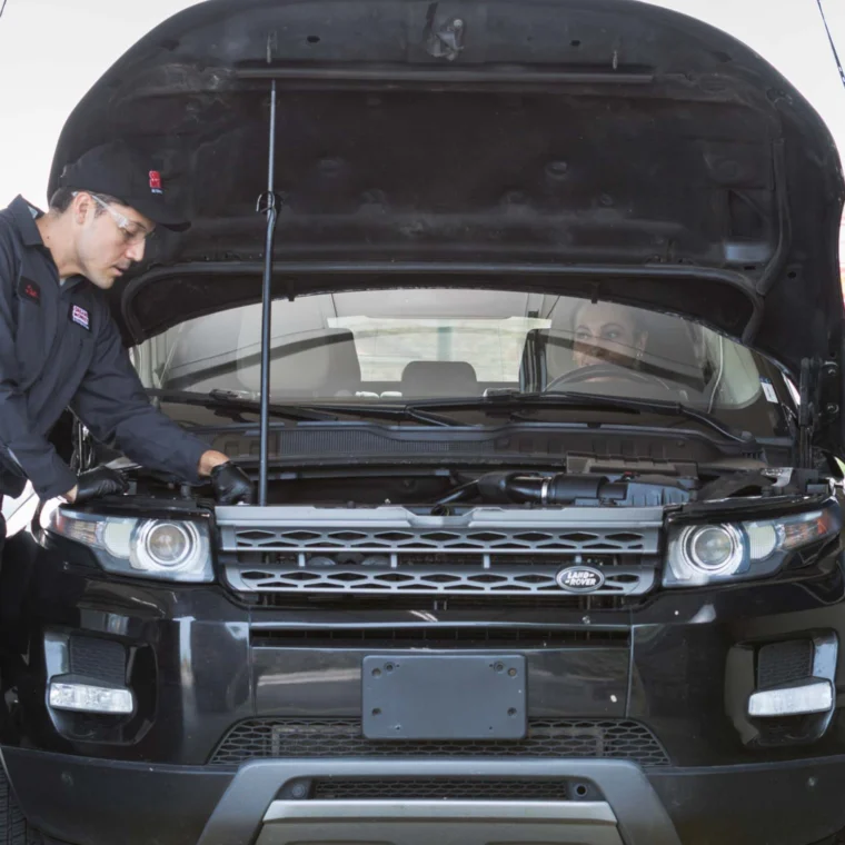 A Grease Monkey mechanic in uniform inspects under the hood of a black SUV with its hood open inside an auto repair shop, preparing for a possible clutch replacement.