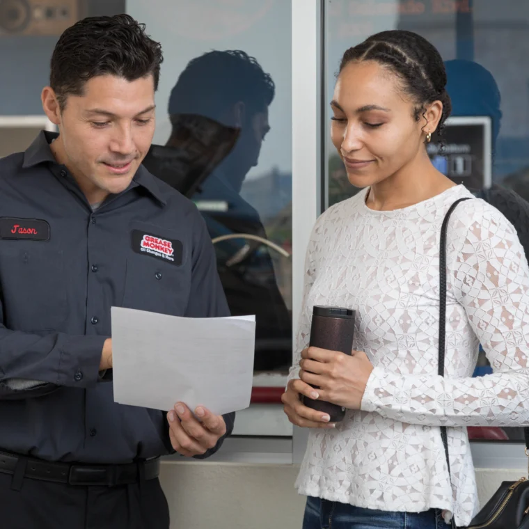 A man in a Grease Monkey uniform showing a document about clutch replacement to a woman holding a travel mug; they are standing near a window.
