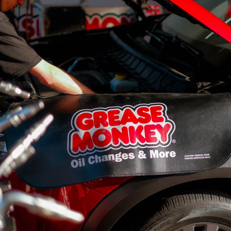 A person performs maintenance under the hood of a red vehicle with a cover labeled "Grease Monkey Oil Changes & More" draped over the front, possibly working on a clutch replacement.