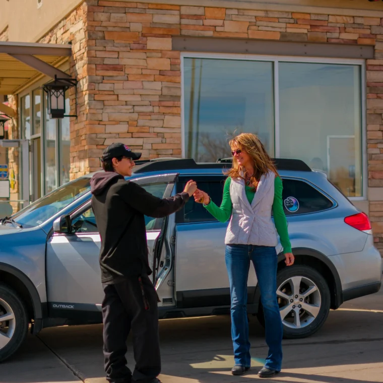 A woman and a man fist bump beside a silver car parked outside a Grease Monkey building with stone walls and large windows, celebrating a successful clutch replacement.