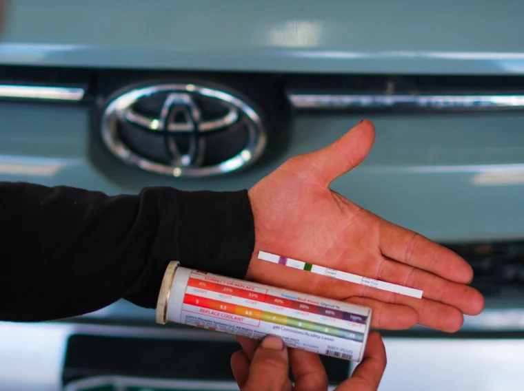 Person holding test strips and a color chart in front of a Toyota, possibly testing automotive fluids under the hood as part of a cooling system repair.