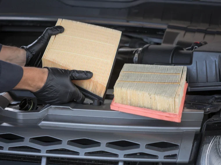 A person wearing black gloves is replacing a dirty car air filter with a clean one under the hood of a vehicle, demonstrating the importance of regular Filter Services for optimal engine performance.