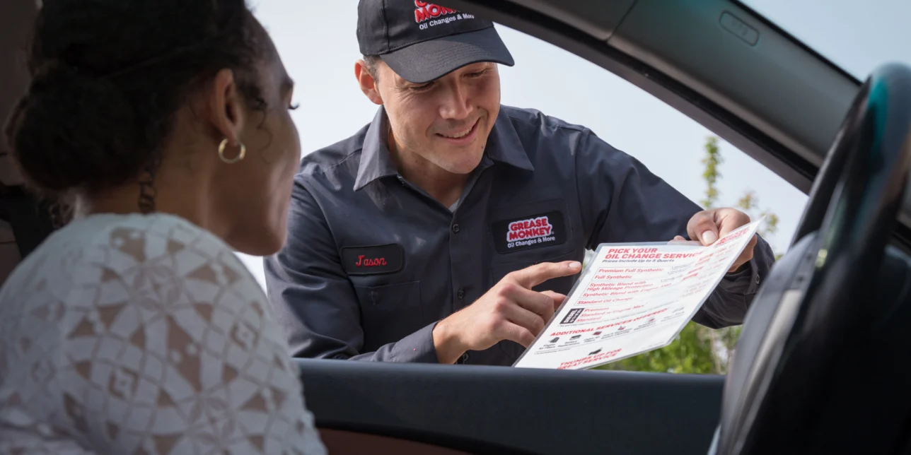 A car service worker shows a printed checklist to a woman sitting in the driver's seat of her car during an outdoor consultation.