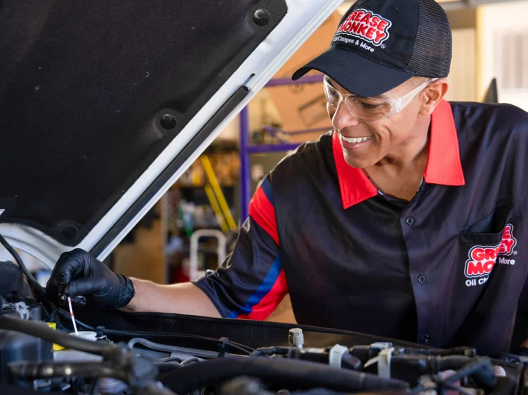 A mechanic wearing safety glasses and gloves checks the oil level of a car engine with a dipstick in an auto repair shop, ensuring proper maintenance as part of the shop’s comprehensive Fluid Exchange Services.