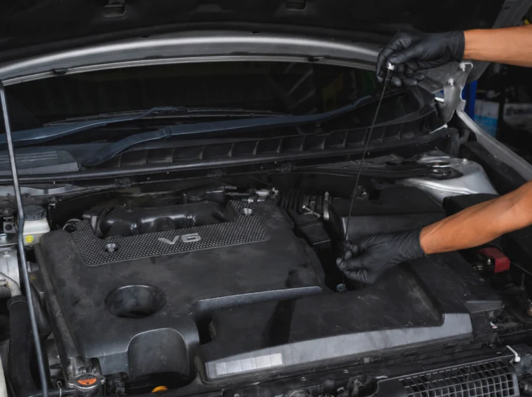 Person wearing black gloves checking a car engine's oil level with a dipstick; the engine cover is labeled "V6." This careful inspection highlights the importance of regular Fluid Exchange Services in maintaining optimal vehicle performance.