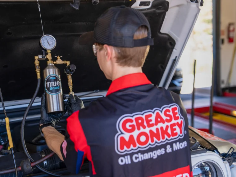 A mechanic wearing a "Grease Monkey" shirt and cap checks engine pressure with a gauge under the hood of a vehicle in an auto shop, ensuring top-quality Engine Services for every customer.
