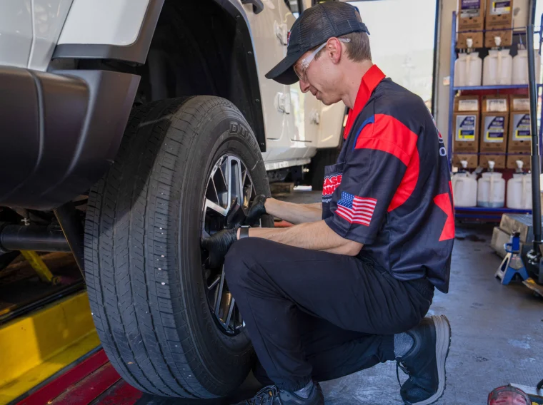 Mechanic wearing gloves and uniform kneels beside a white vehicle, inspecting or working on the front tire inside an auto service garage, providing expert Tire Services.