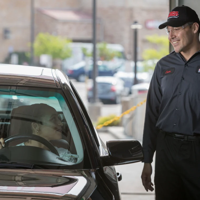 A person in a car speaks with a smiling service worker in uniform standing outside a building entrance, ready to assist with expert car detailing.