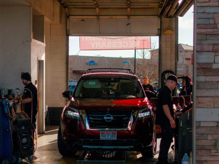 A Nissan SUV is parked inside a car service garage as Grease Monkey technicians work around it, with equipment and tools visible on the sides, providing professional car wash services.