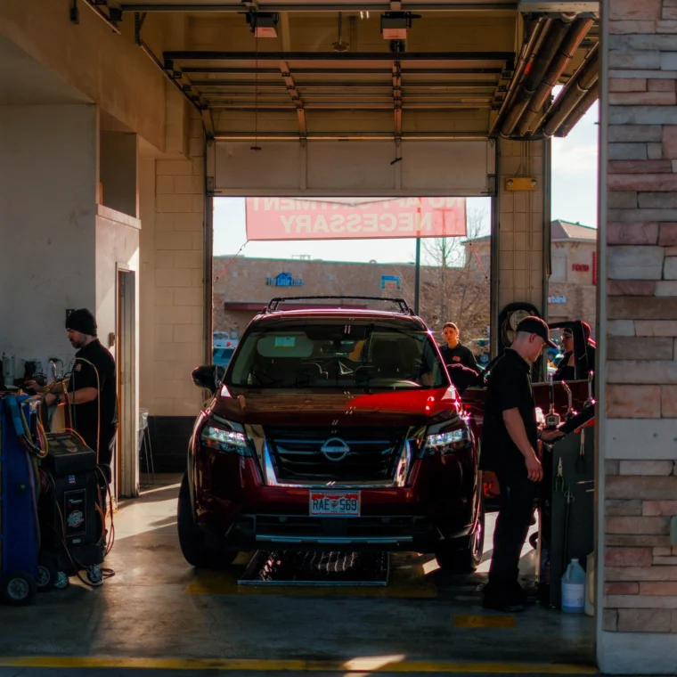 A Nissan SUV is parked inside a car service garage as Grease Monkey technicians work around it, with equipment and tools visible on the sides, providing professional car wash services.