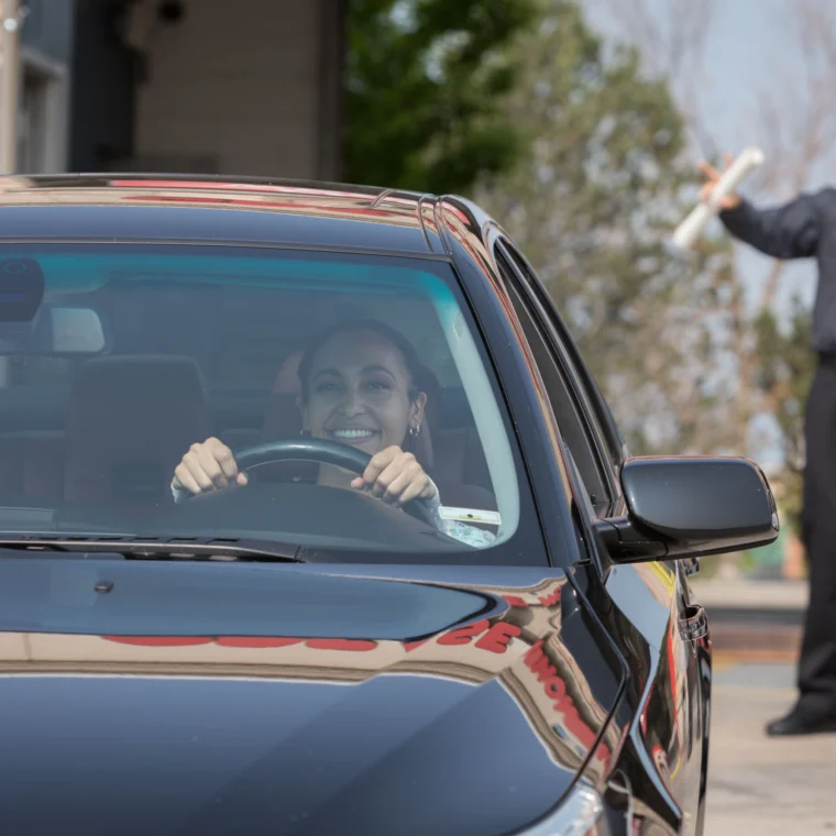 A woman smiles while sitting in the driver’s seat of a black car at a drive-thru Car Wash, as a uniformed worker stands outside holding a rolled-up document.