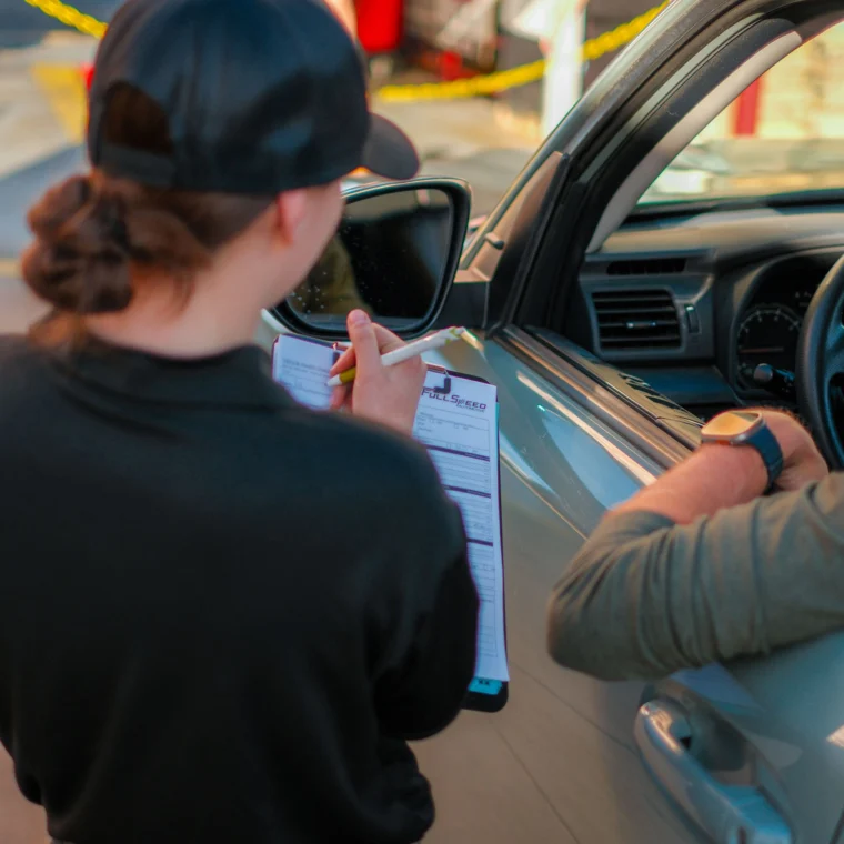 A worker holding a clipboard speaks with a person in a car at a service station, while other employees handle car cleaning tasks in the background near caution tape.