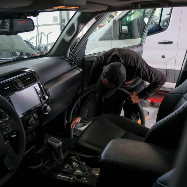 A person in a uniform is vacuuming the interior floor of a parked car at an auto detailing facility, showcasing professional car wash services.