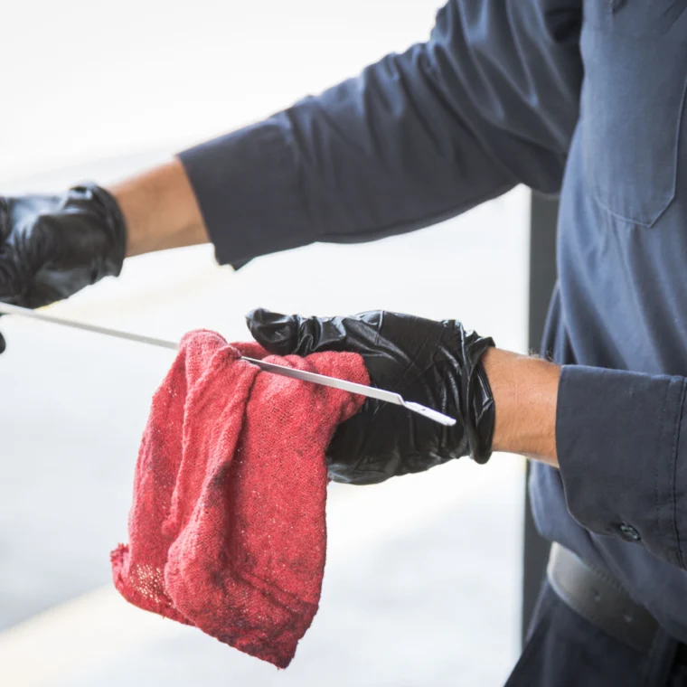 A person wearing black gloves wipes an engine oil dipstick with a red cloth, likely performing vehicle maintenance as part of routine engine services.
