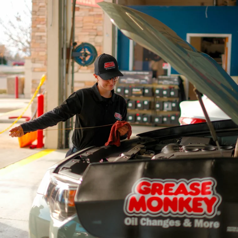 A mechanic in a Grease Monkey uniform checks the oil level under the hood of a vehicle, showcasing expert Engine Services inside an auto service garage.