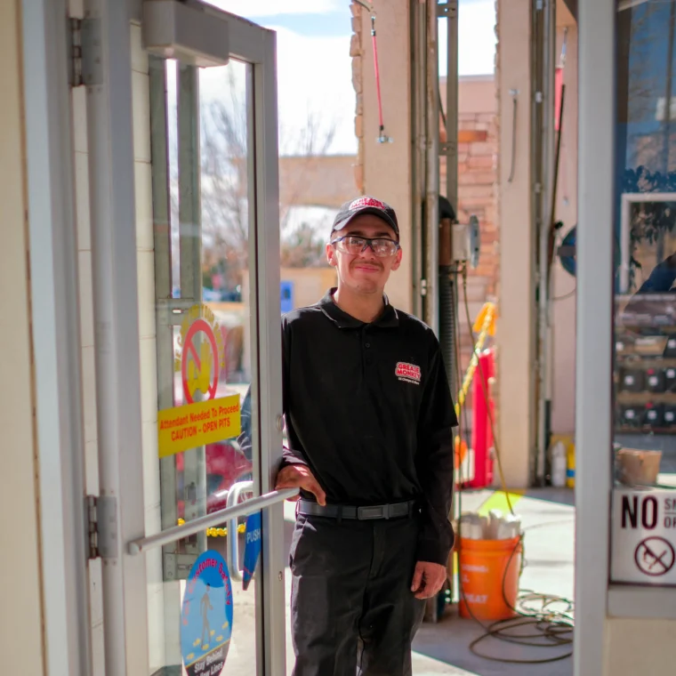 A man in a Grease Monkey uniform and hat stands at an open glass door, smiling, with a service area visible in the background—highlighting expert Engine Services offered on site.