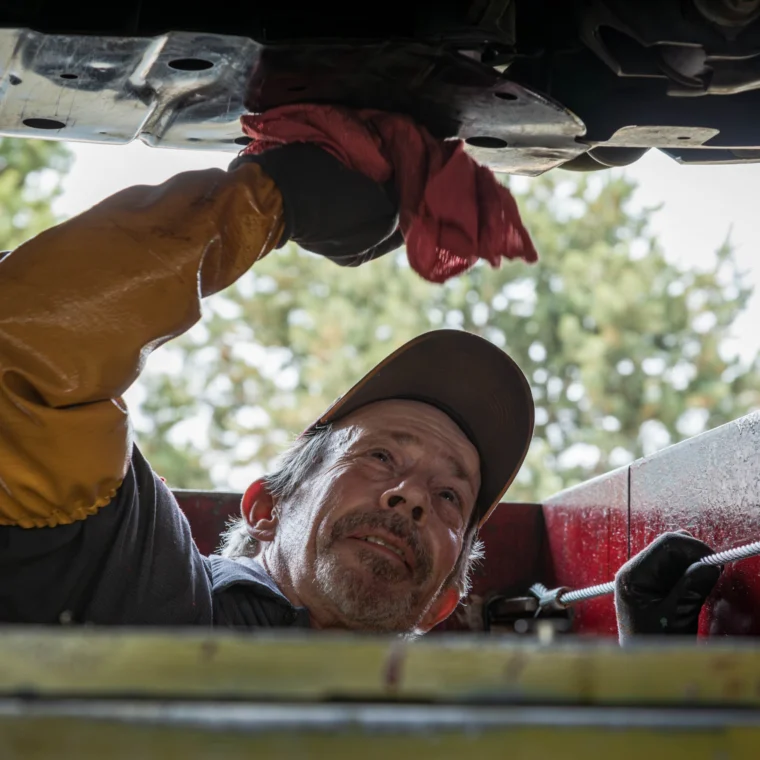 A mechanic wearing gloves and a cap wipes the underside of an EV with a red cloth while performing EV maintenance in a garage.